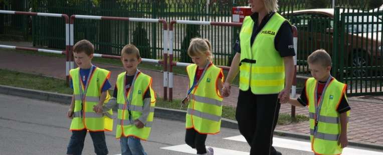Dzieci oraz policjantka w kamizelkach odblaskowych, na pasach